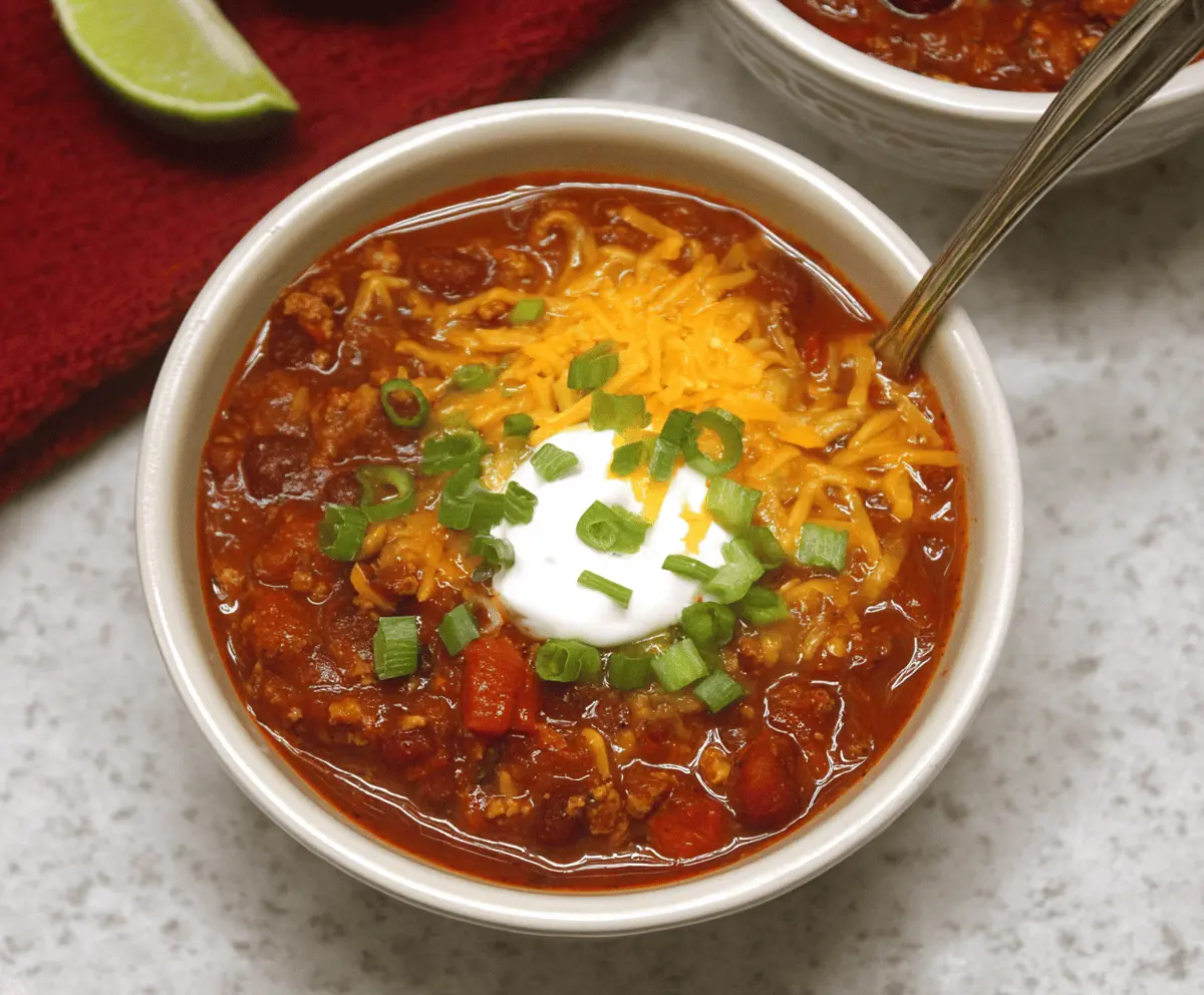 A steaming bowl of gluten-free chili topped with shredded cheese, fresh cilantro, and diced onions, served with cornbread on the side.