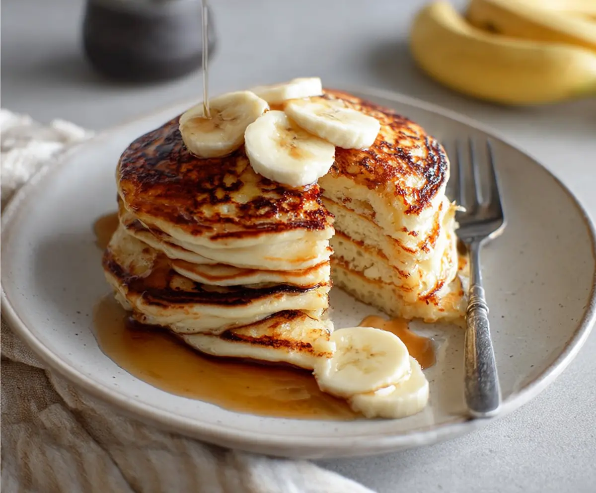 Delicious banana yogurt pancakes stacked on a plate, topped with fresh banana slices and syrup.