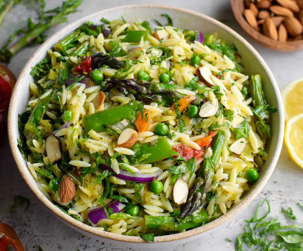 Colorful spring orzo pasta salad with fresh vegetables and herbs in a clear glass bowl.